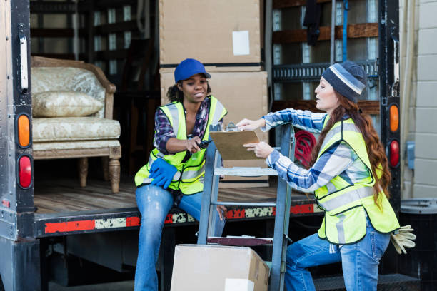 Two multi-ethnic female workers in their 30s and 40s at the back of a truck, one holding a clipboard. The women are wearing reflective vests and jeans. They are moving merchandise for a furniture store.